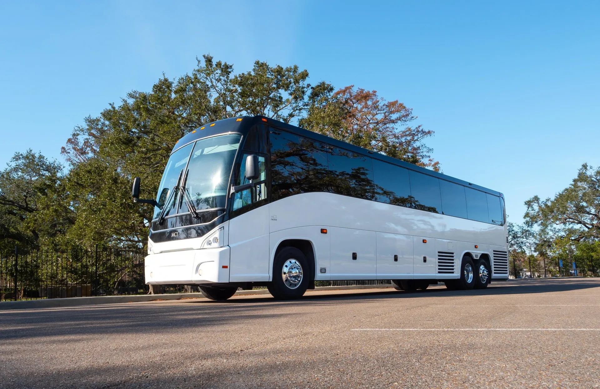 A sleek white MCI motorcoach bus parked on a sunny day in an open lot with trees and a clear blue sky in the background.