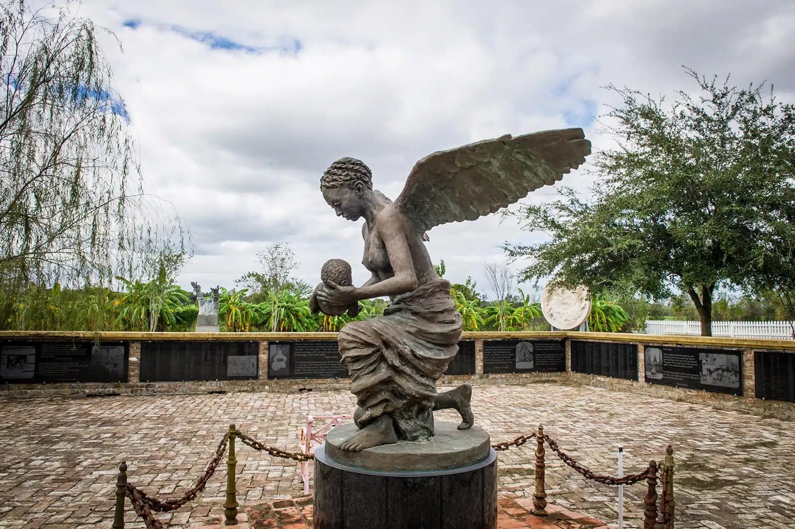 A bronze statue of an angel kneeling while holding a baby, set on a circular pedestal surrounded by a brick courtyard and informational plaques, with trees and a cloudy sky in the background.