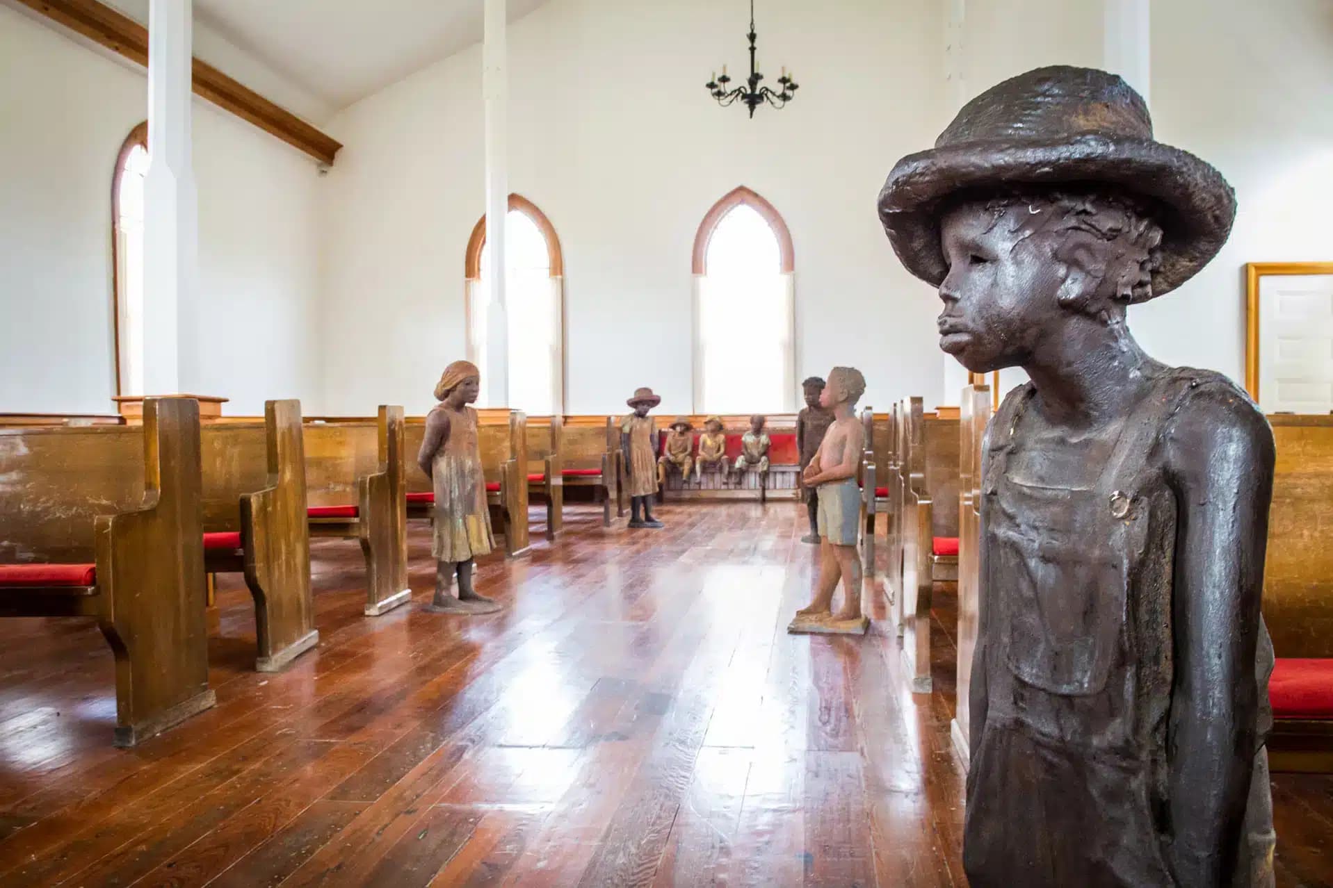 The interior of a church featuring wooden pews and statues of children in historic attire standing throughout the space, with arched windows and wooden flooring.