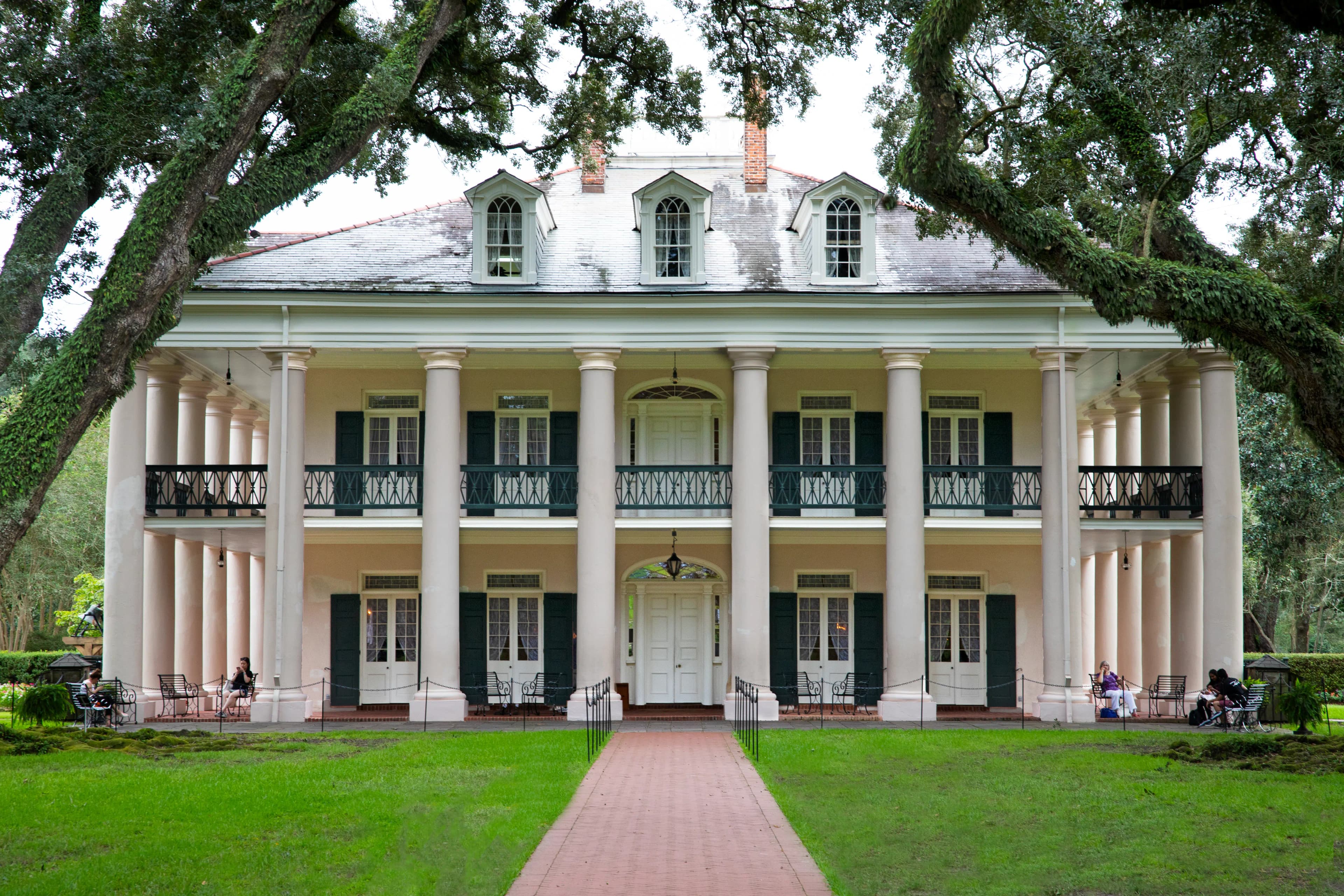 The front view of a grand plantation house with tall white columns, green shutters, and a red brick pathway leading to the main entrance, surrounded by lush trees and greenery.