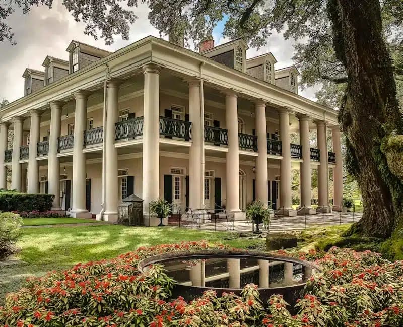 A grand plantation house with tall white columns, a wraparound balcony, and manicured gardens featuring vibrant red flowers and a reflective fountain in the foreground.