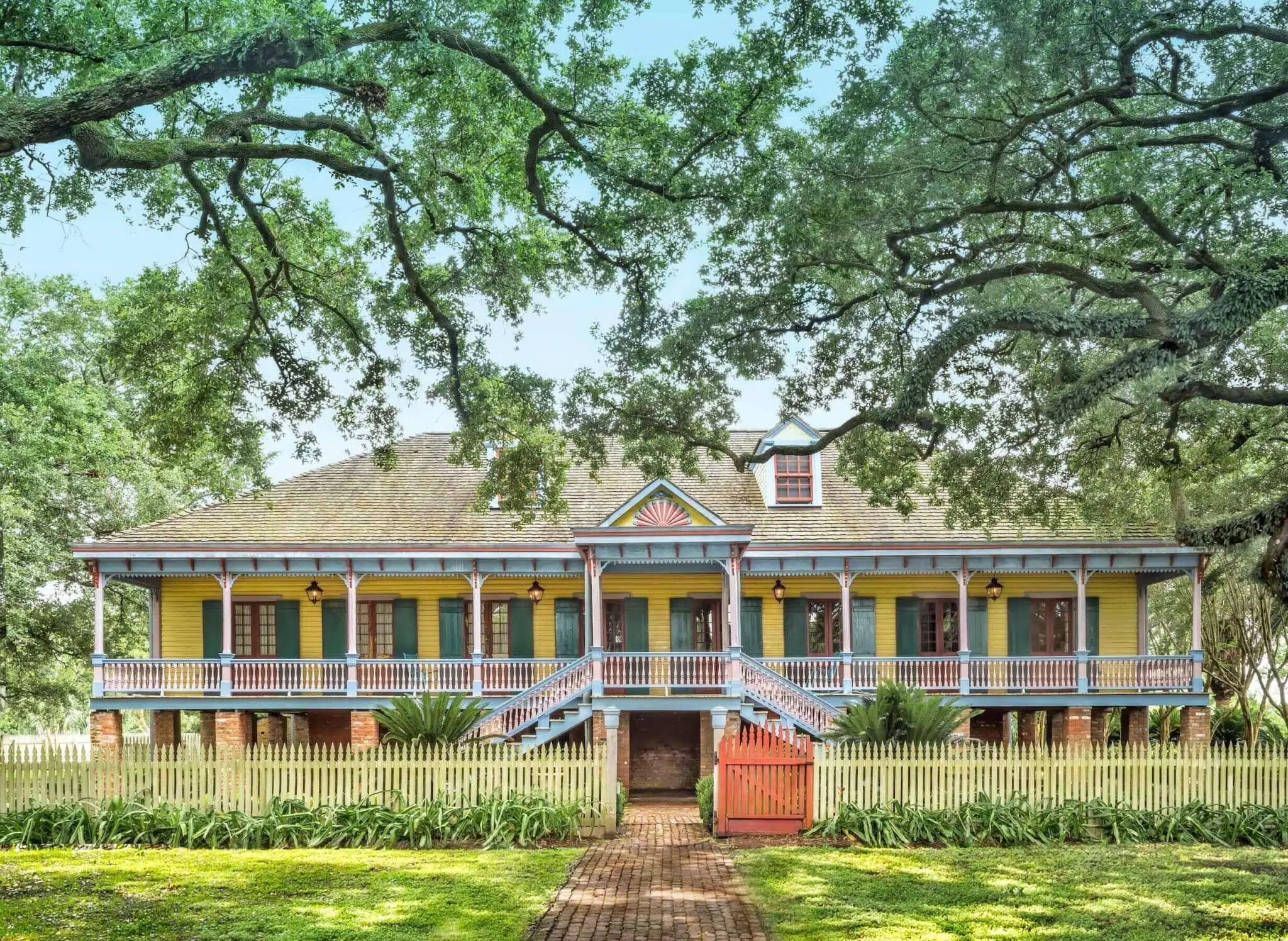 A vibrant yellow plantation house with green shutters, blue trim, and twin staircases, surrounded by a white picket fence, lush greenery, and shaded by large oak trees.