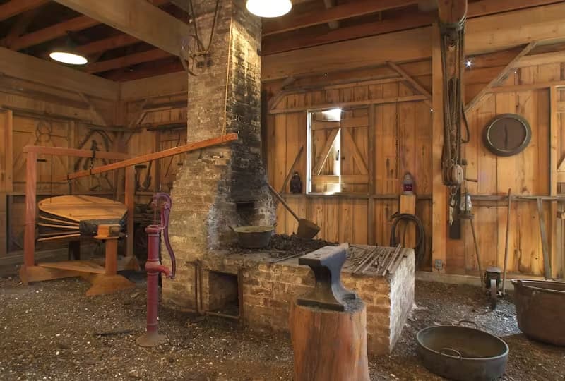 The interior of a historic blacksmith shop featuring a brick forge, an anvil on a wooden block, a red water pump, and various tools hanging on wooden walls.