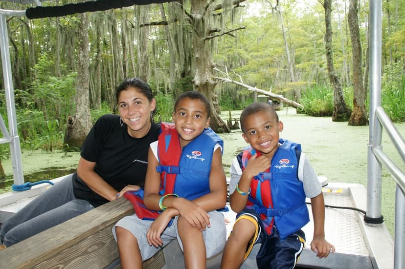 Woman and two young boys in life jackets smile while seated on a swamp tour boat surrounded by lush cypress trees.