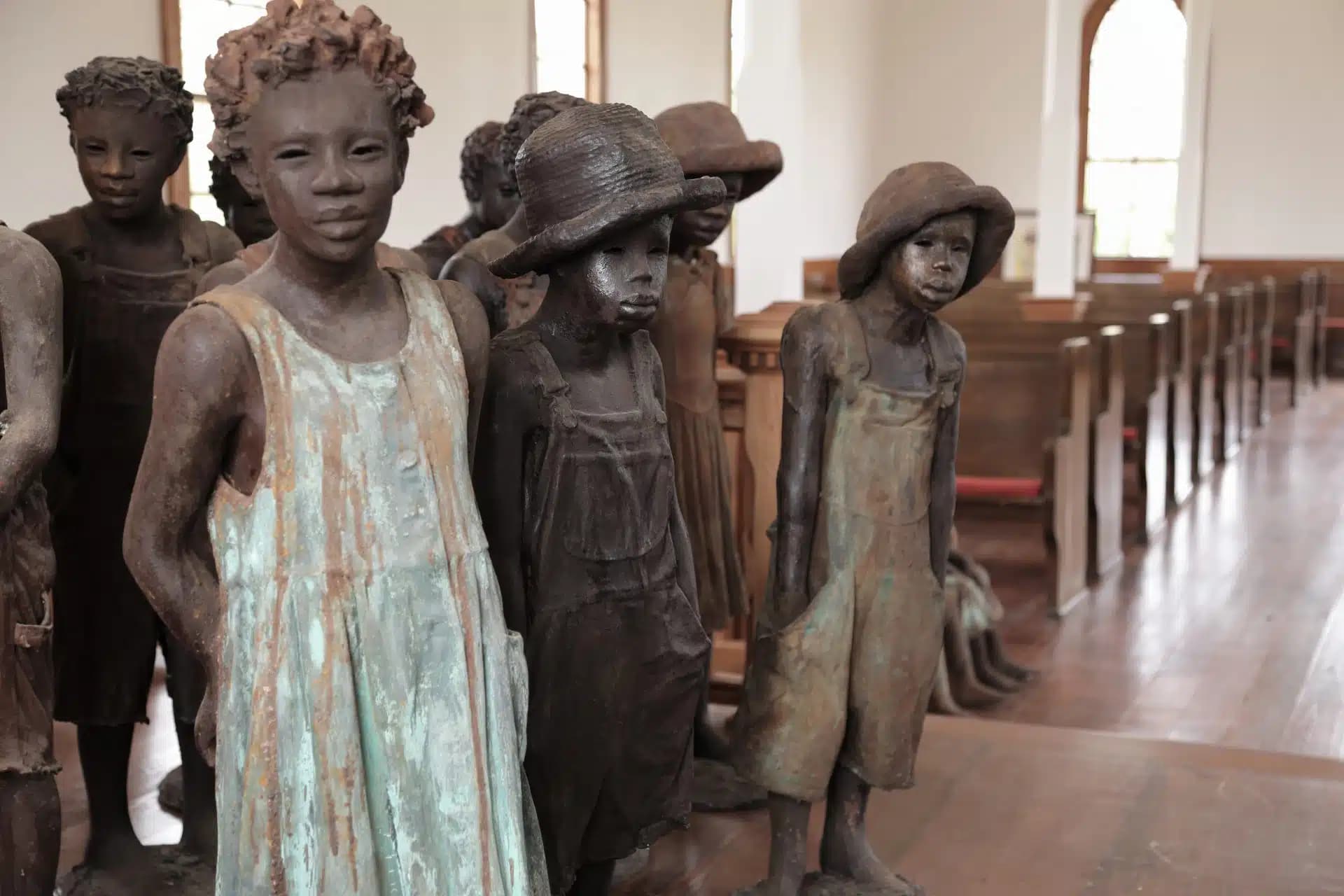 Close-up of statues depicting children in historic attire, including dresses and overalls, standing inside a church with wooden pews and arched windows in the background