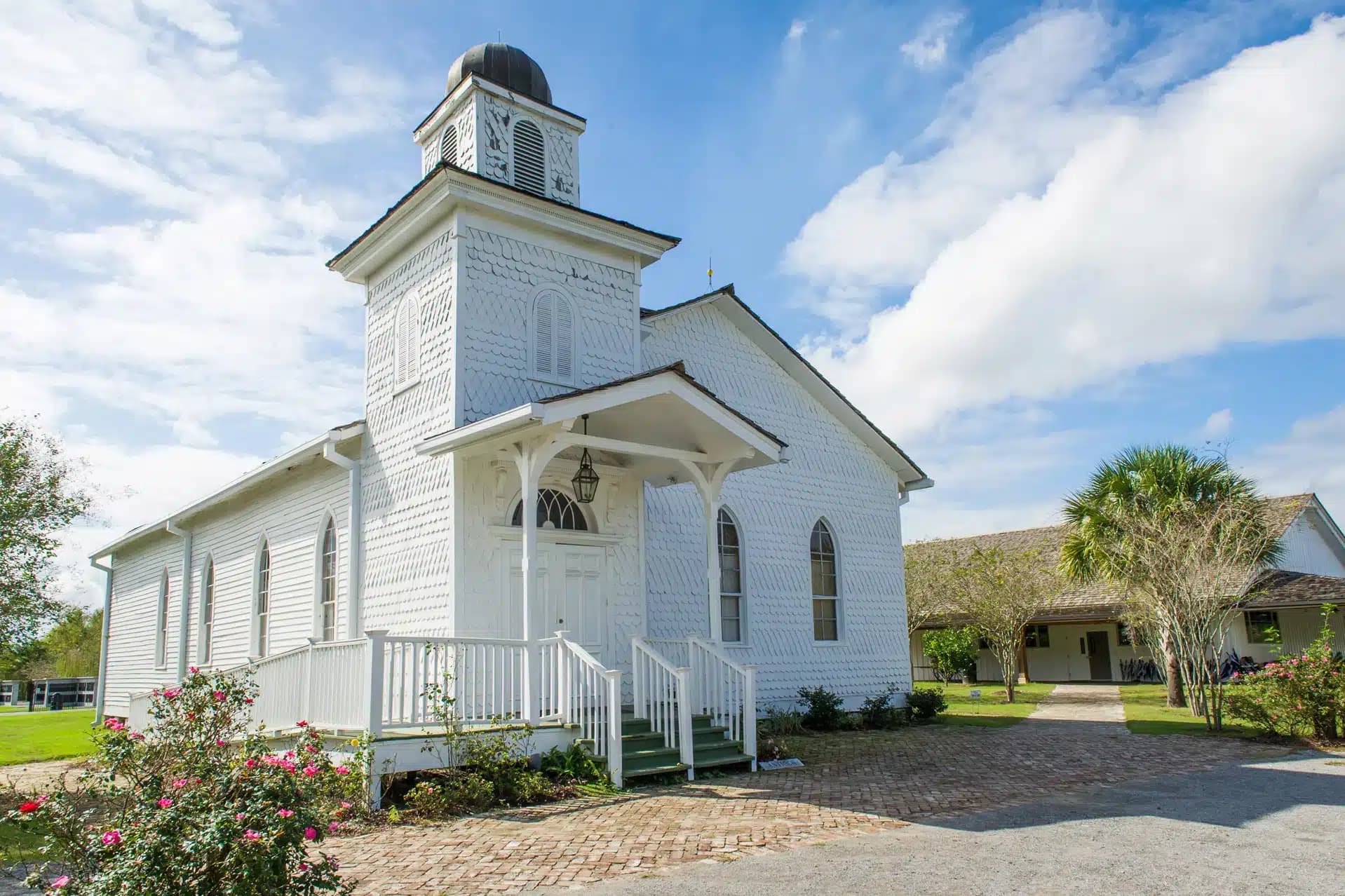 A white wooden church with arched windows, a small steeple, and a front porch, surrounded by rose bushes and a brick walkway under a bright blue sky.