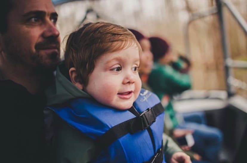 Young child wearing a blue life jacket smiles while sitting on a boat, held by a man looking into the distance.