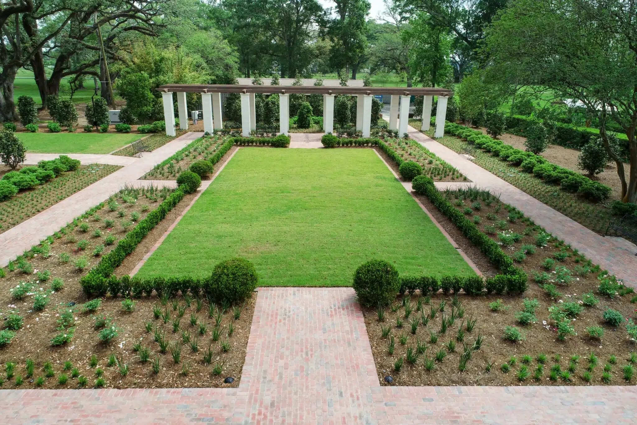 A symmetrical garden with a large rectangular lawn bordered by hedges, flower beds, and brick pathways, leading to a pergola with white columns in the background.