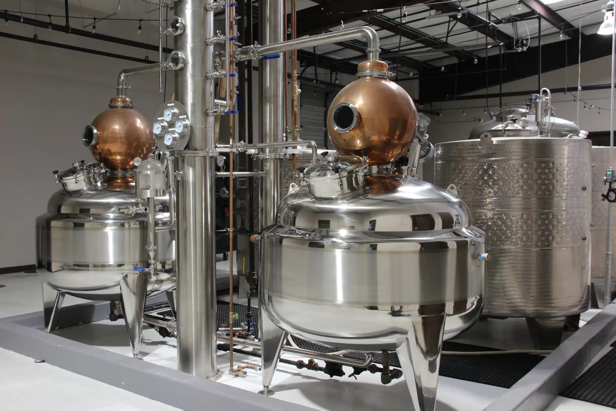 A distillery setup featuring shiny stainless steel and copper stills with connected pipes and gauges, set in an industrial space with exposed beams and polished flooring.