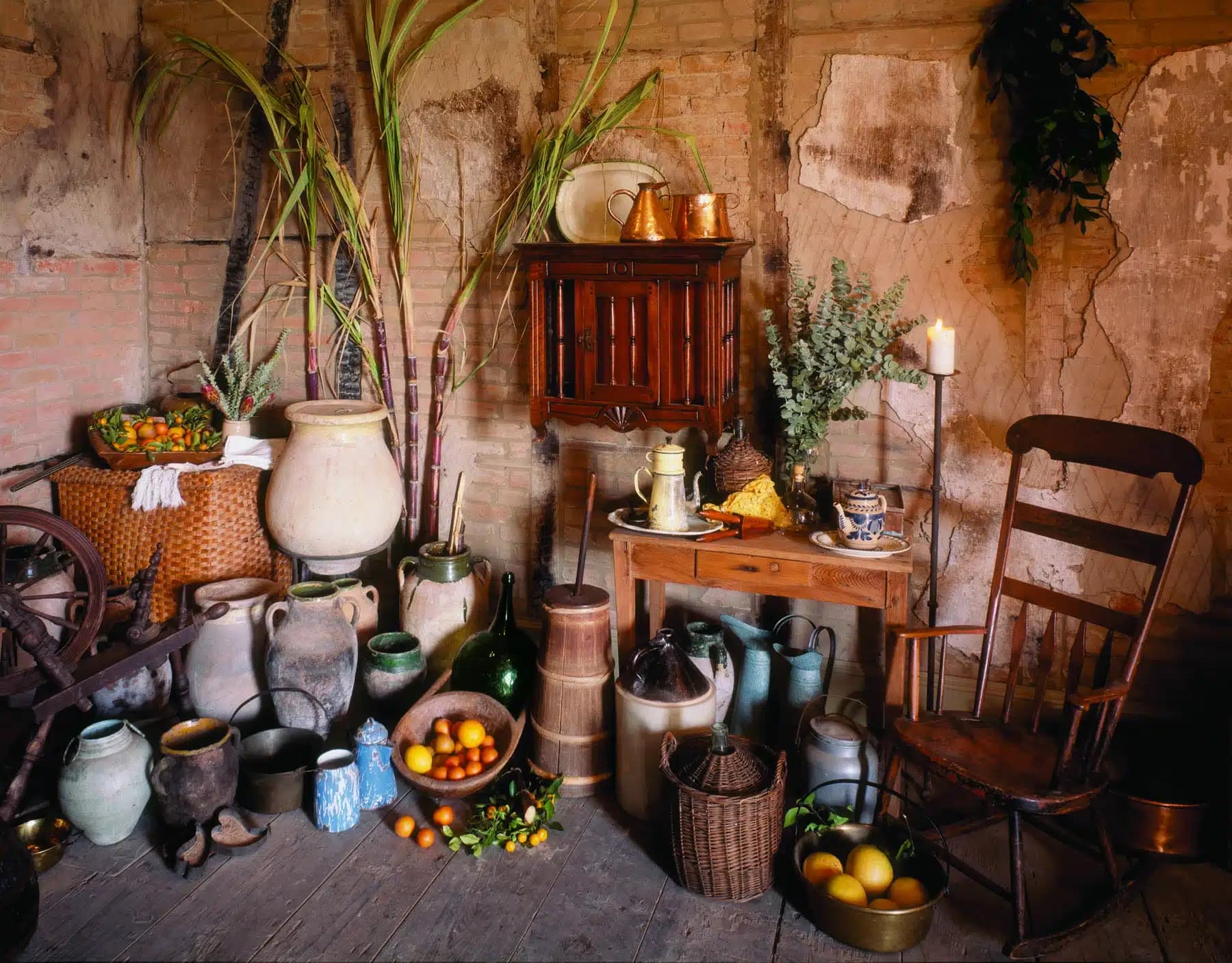 A rustic interior scene featuring earthenware pots, wicker baskets, sugarcane stalks, fresh fruit, and a wooden rocking chair, with a small table holding a candle, dishes, and greenery against a weathered brick wall.