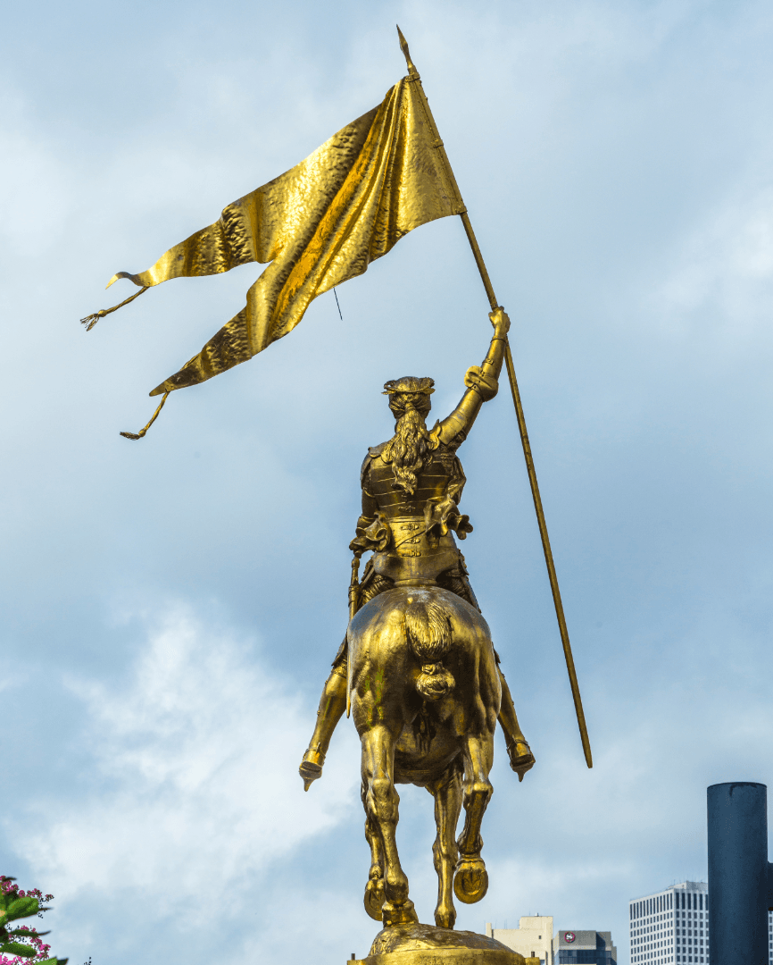 Golden equestrian statue of Joan of Arc holding a flag, set against a cloudy sky with glimpses of buildings in the background, located in the New Orleans French Quarter.