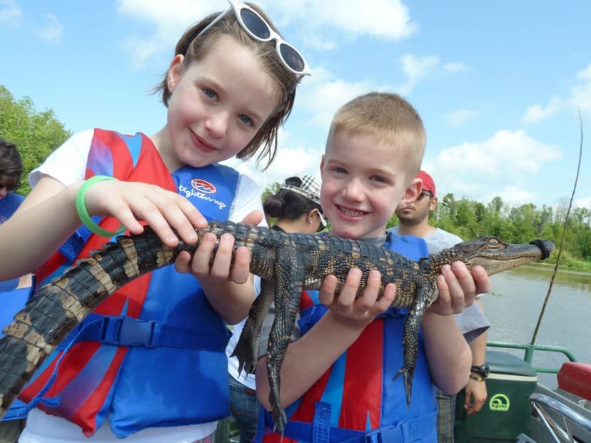 Two children in life jackets smile while holding a baby alligator on a sunny day by a swamp.