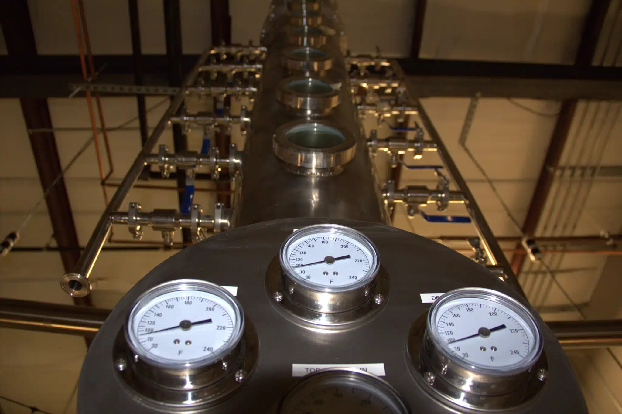 Close-up of a tall distillation column with multiple glass viewing ports, surrounded by connected pipes and gauges, in an industrial distillery setting.