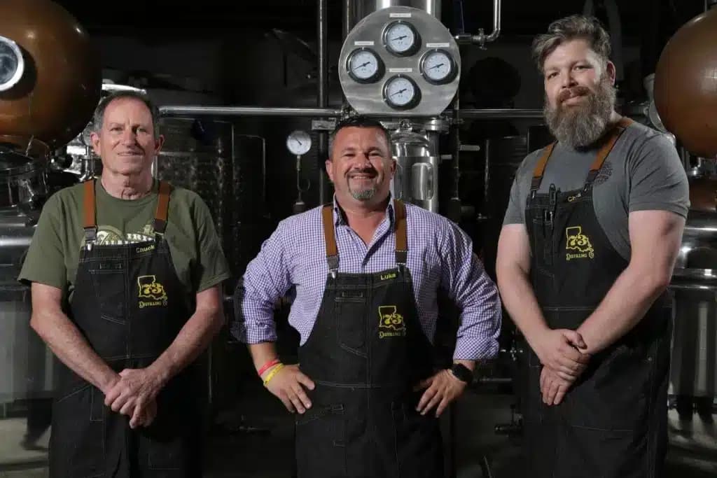 Three distillers standing in front of a distillery setup with copper and stainless steel stills, wearing black aprons with a distillery logo, smiling confidently.