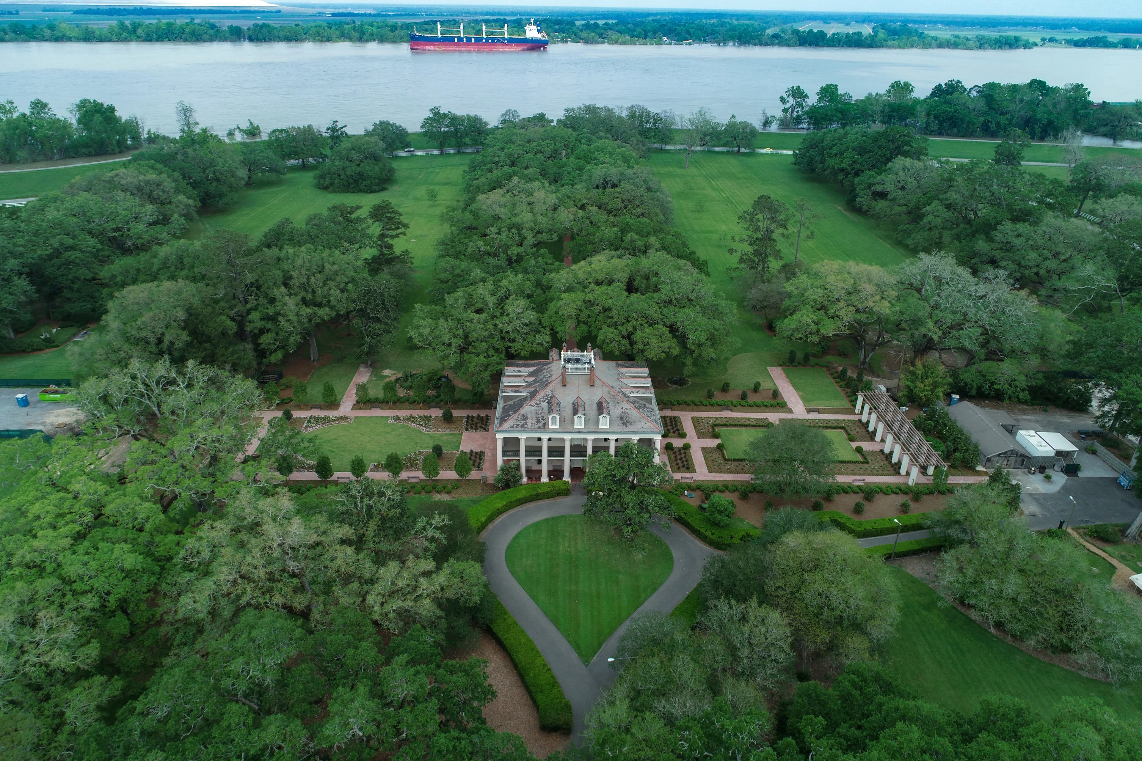 Aerial view of a plantation estate featuring a large house with white columns, symmetrical gardens, and oak tree-lined pathways, with a river and cargo ship in the background.