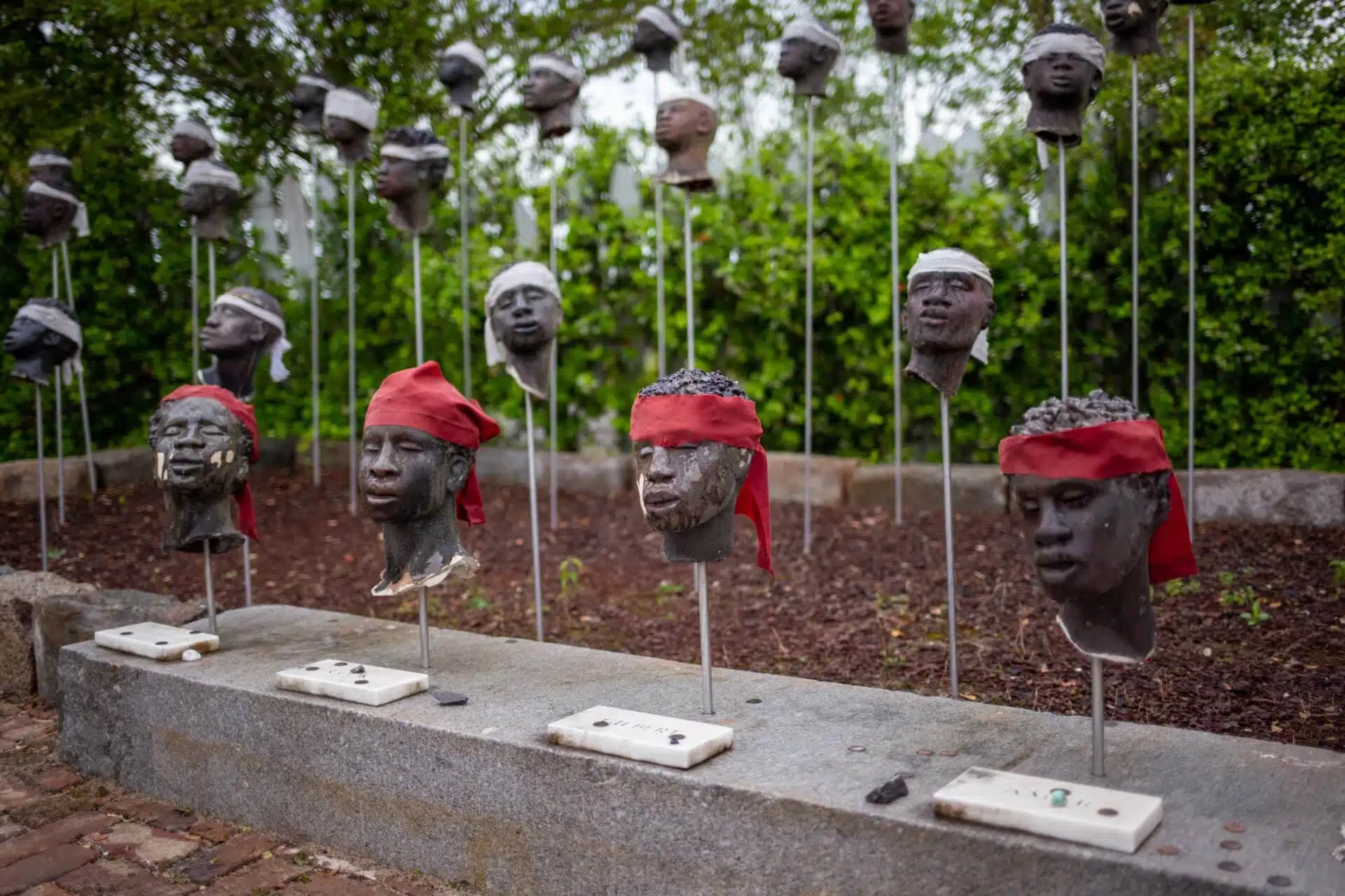 A memorial featuring sculpted heads mounted on metal rods, some with red or white cloth headbands, set in a garden with brick paths and greenery in the background.