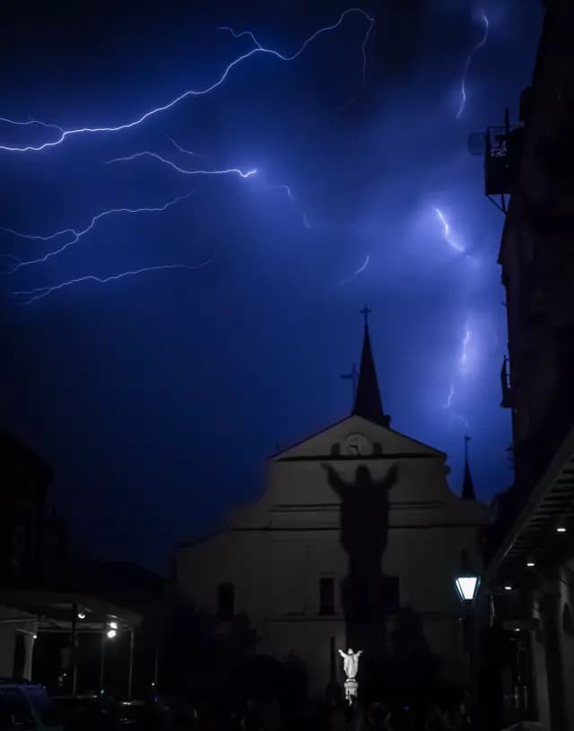 Lightning illuminates a church with a statue casting an ominous shadow on the building under a dark blue sky.