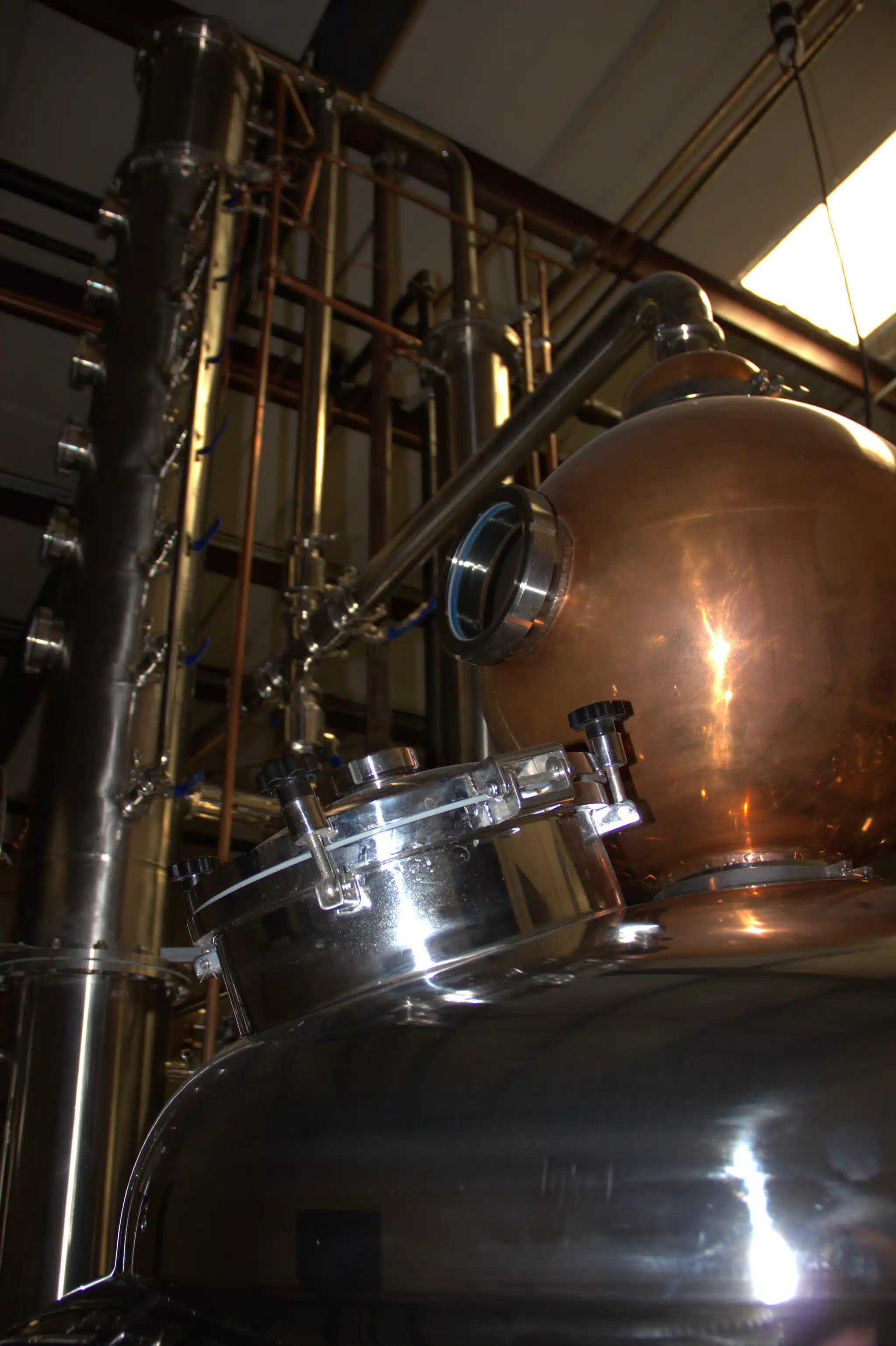 Close-up of a shiny copper and stainless steel distillation apparatus with connected pipes and columns in an industrial distillery setting.