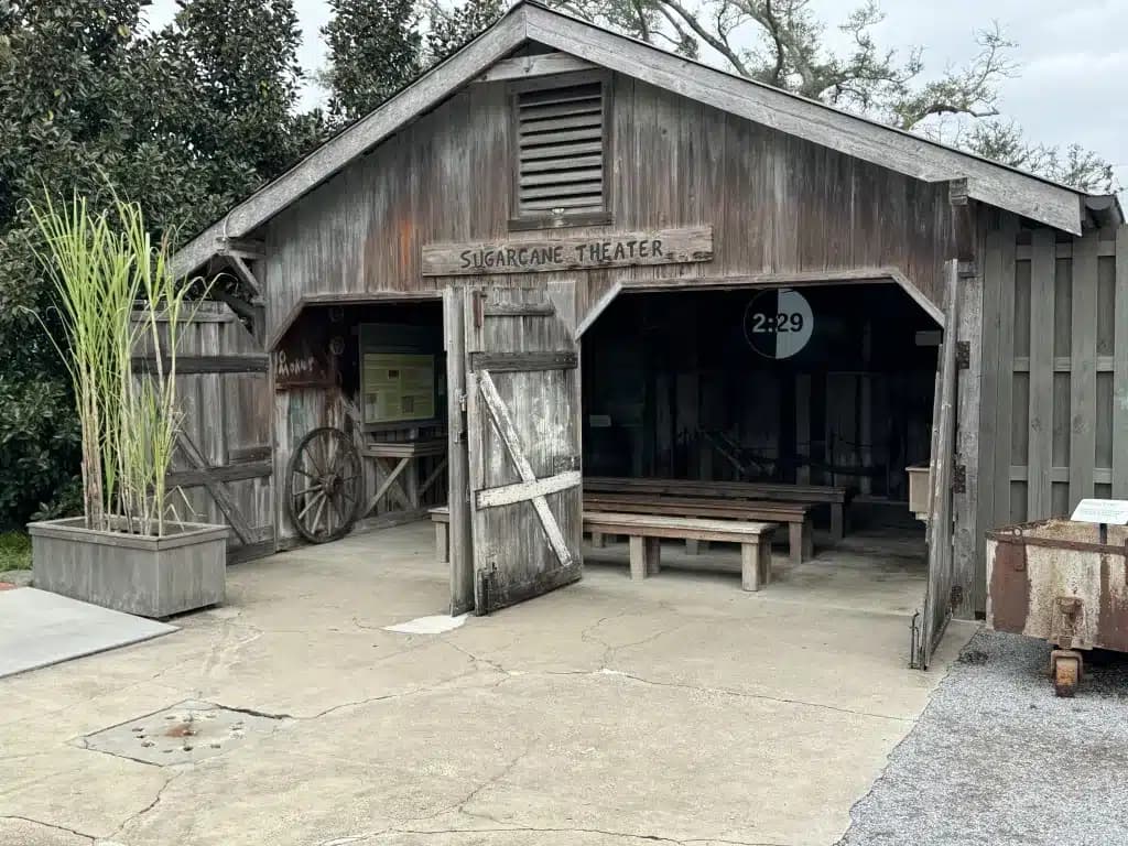 A rustic wooden building labeled 'Sugarcane Theater,' featuring open barn doors, wooden benches inside, and sugarcane plants in planters outside.