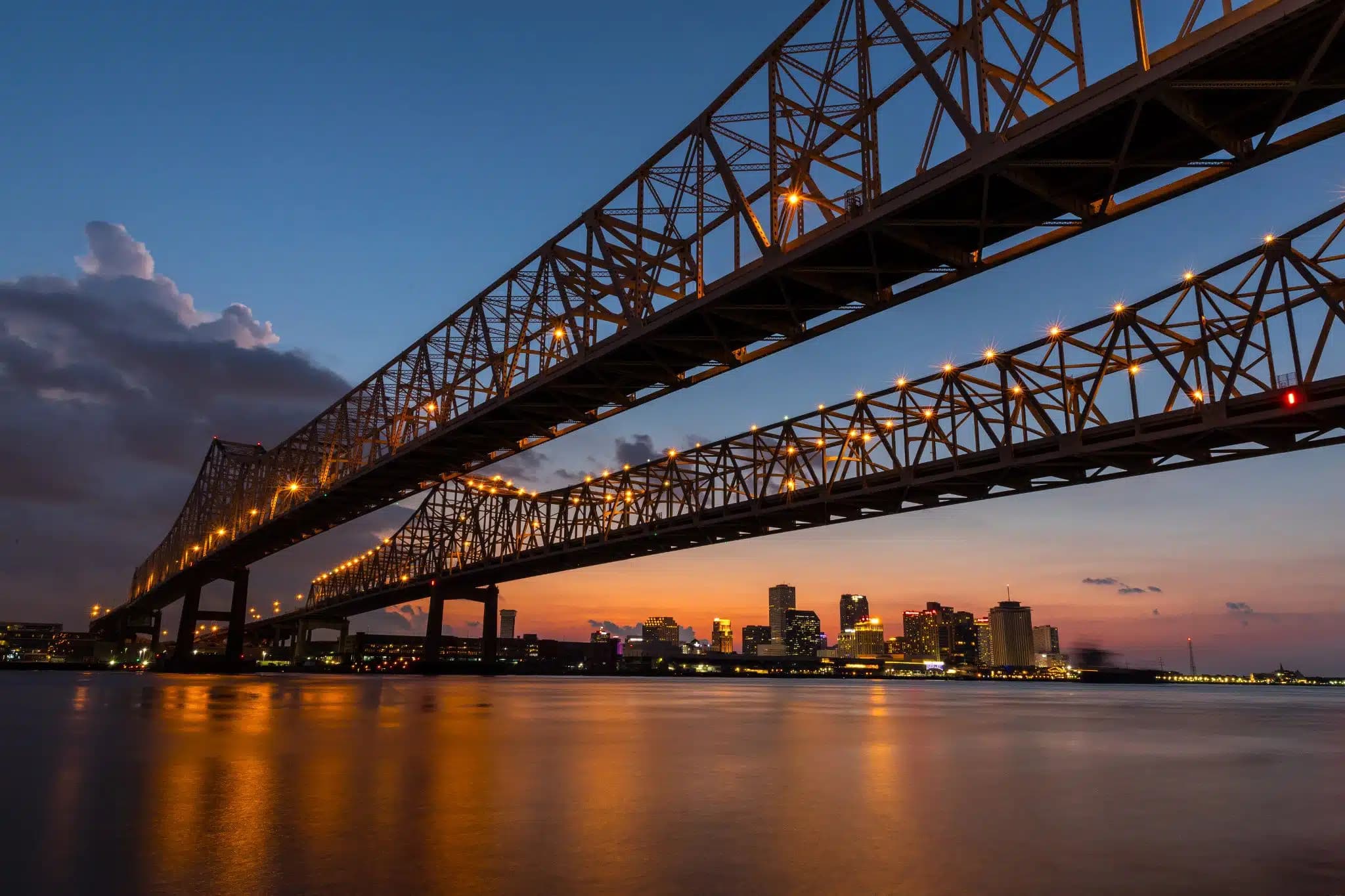 Lit Crescent City Connection bridges over the Mississippi River with the New Orleans skyline at sunset.