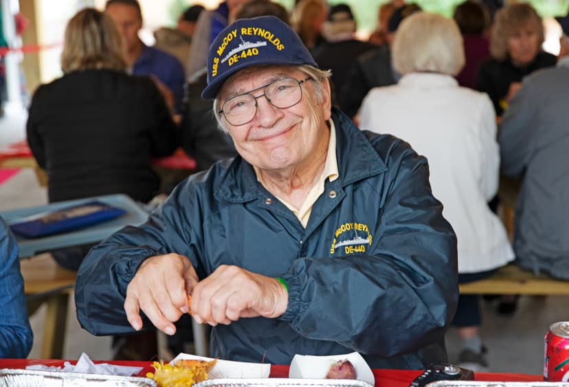 Elderly man wearing a USS McCoy Reynolds DE-440 hat and jacket smiles while seated at a picnic table, preparing to eat.