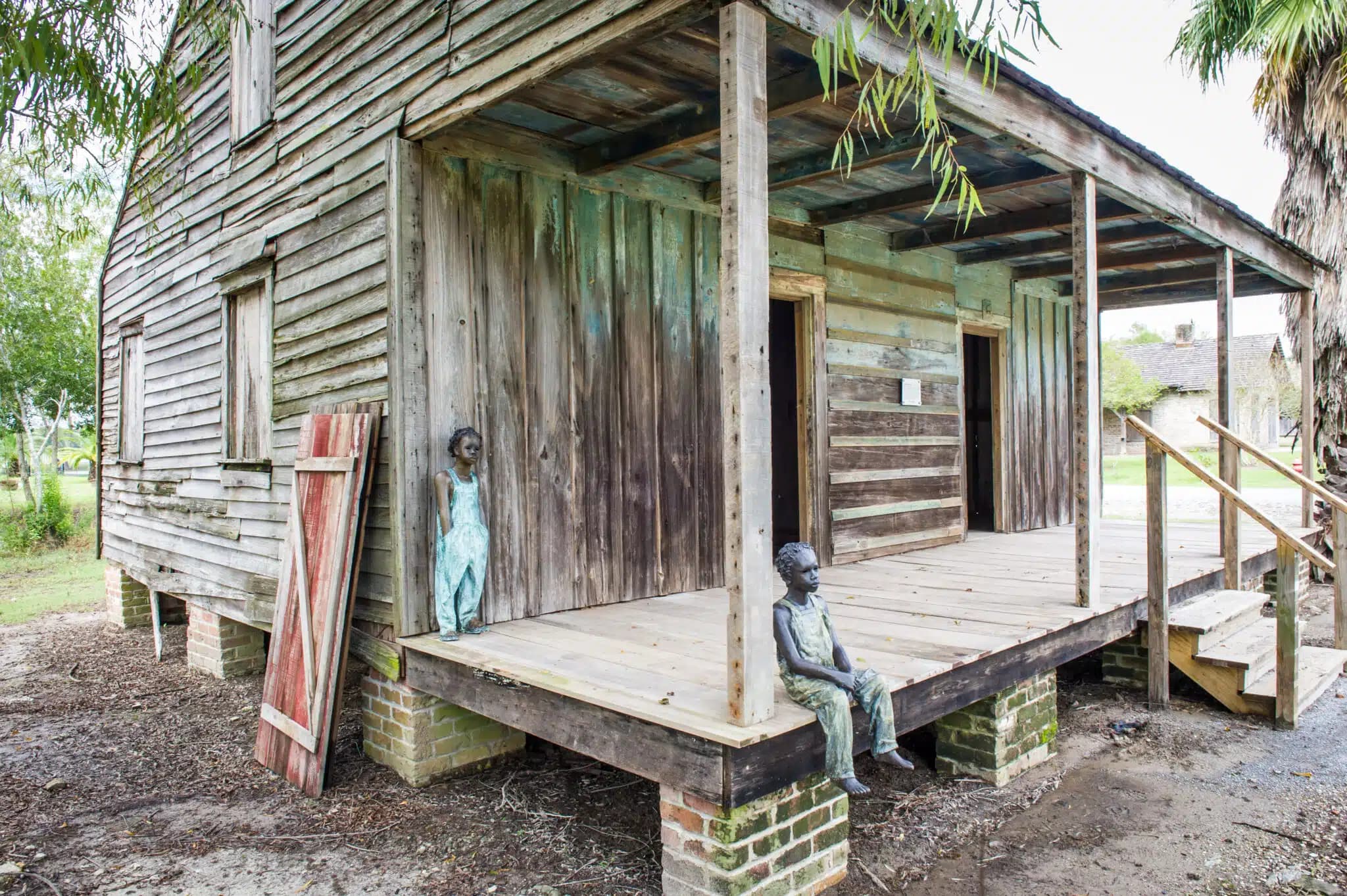 A weathered wooden slave cabin with a front porch on brick pillars, featuring two statues of children in worn clothing, set against a grassy yard with trees in the background.